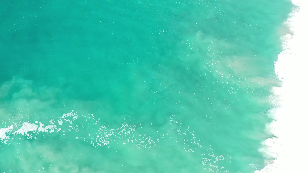 Overhead Los Cabos ocean frame with teal water, small foam flecks across the middle, and a bright band of white surf on the right, no visible people.