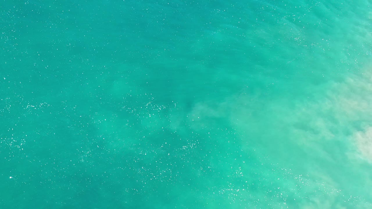 Overhead view of turquoise water in Los Cabos, Mexico, with pale foamy water spreading along the right edge and no visible people.