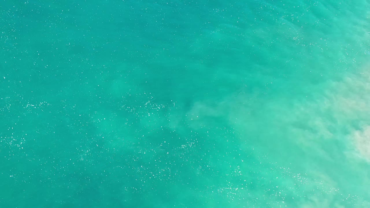Overhead Los Cabos water frame with scattered white flecks on turquoise water and a cloudy foam area crossing the upper right, with no visible people.