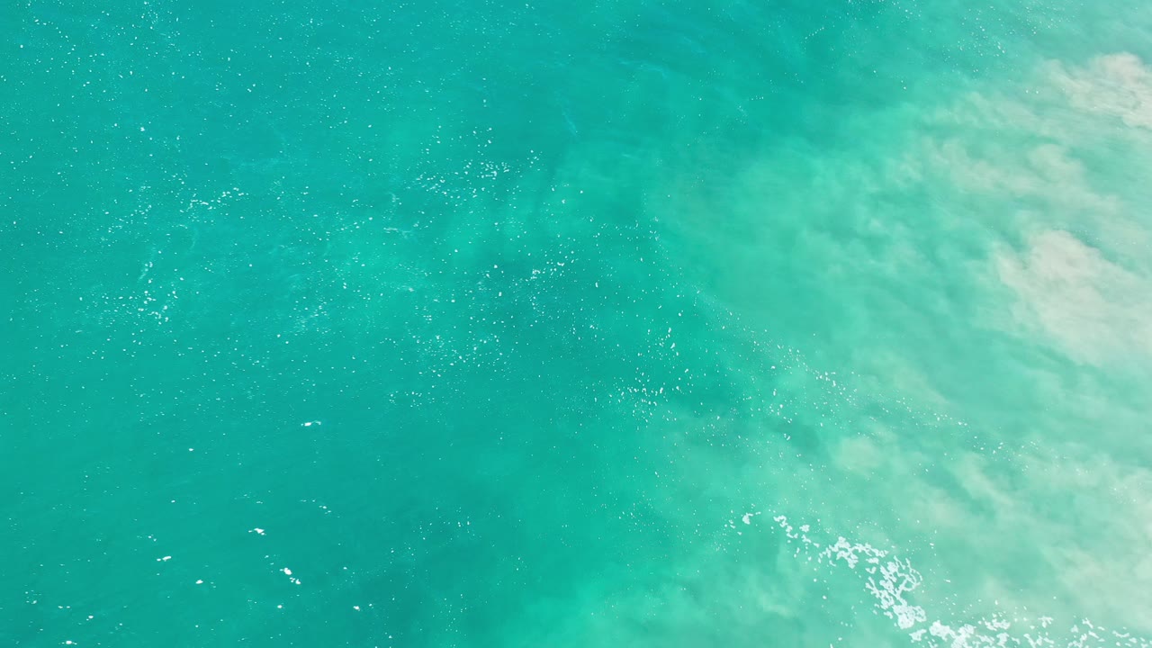 Overhead view of turquoise seawater in Los Cabos, Mexico with pale foam and sandy clouding on the right side, no visible people.