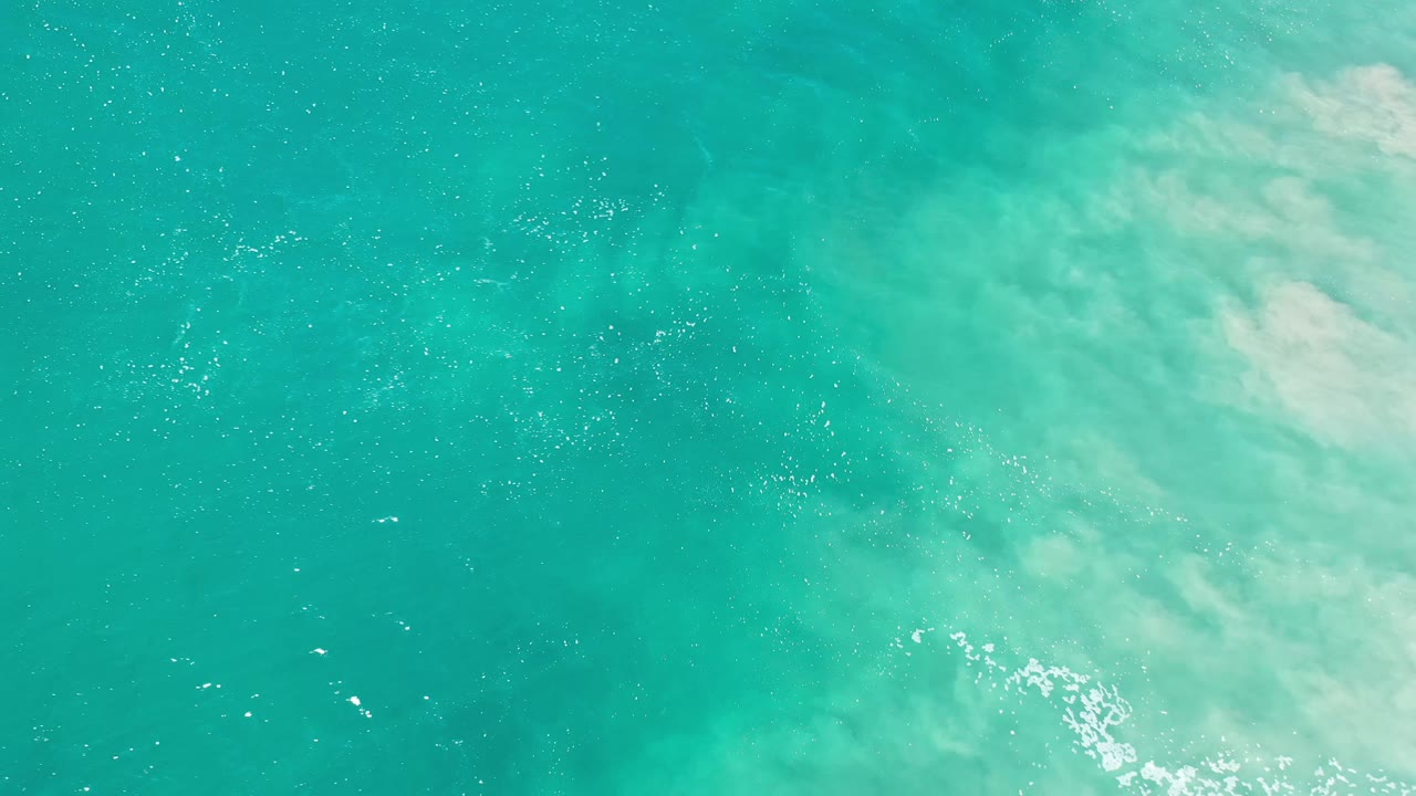 Overhead Los Cabos seawater frame with scattered sun glints, teal color variation, and foam concentrated near the lower right, no visible people.