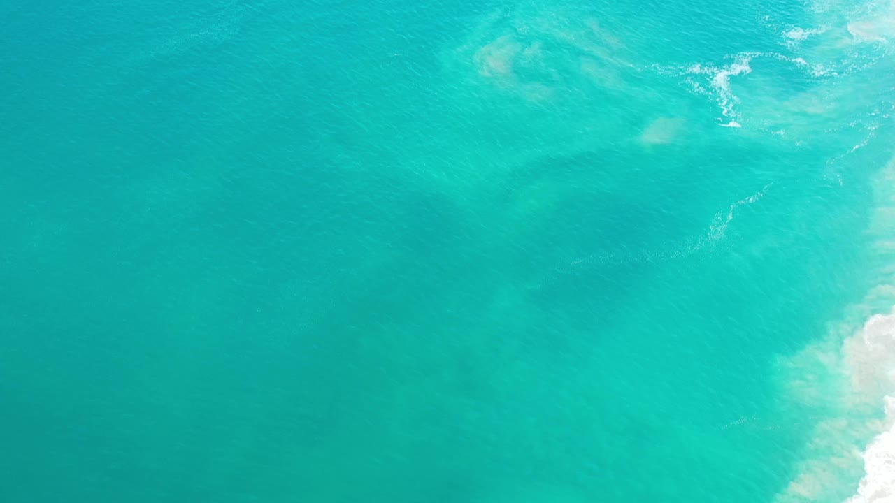 Overhead Los Cabos ocean frame with blue-green water, subtle darker underwater areas, a small surf line near the top right, and no visible people.