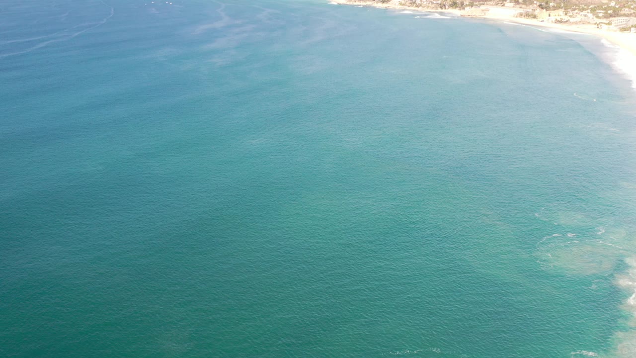 Opening aerial frame over turquoise water in Los Cabos, Mexico, with pale surf at the upper right edge and no visible people.