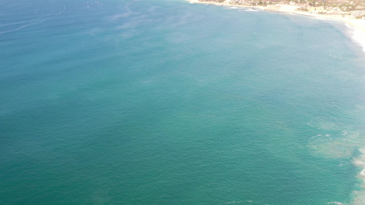 Aerial view in Los Cabos, Mexico, with teal water filling most of the frame and a sunlit strip of beach near the upper right, no visible people.