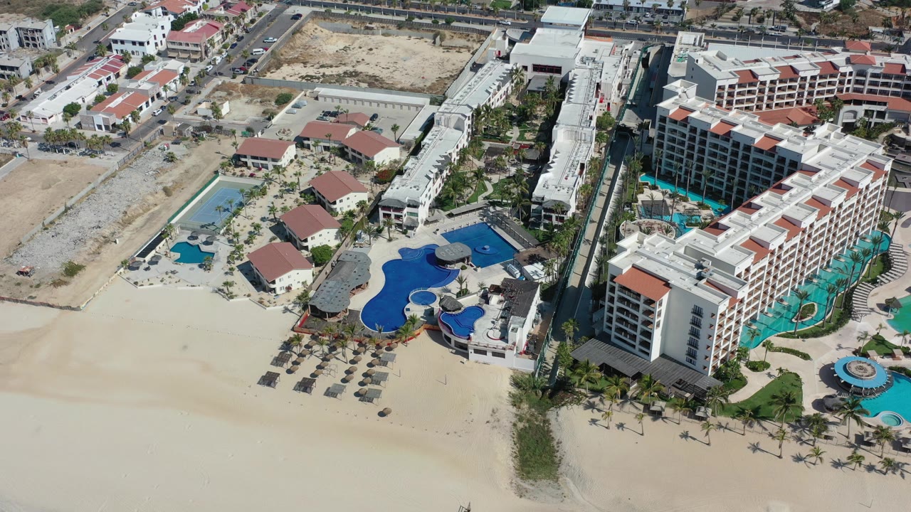 Aerial view of beachfront resort buildings, pools, sand, and umbrellas.