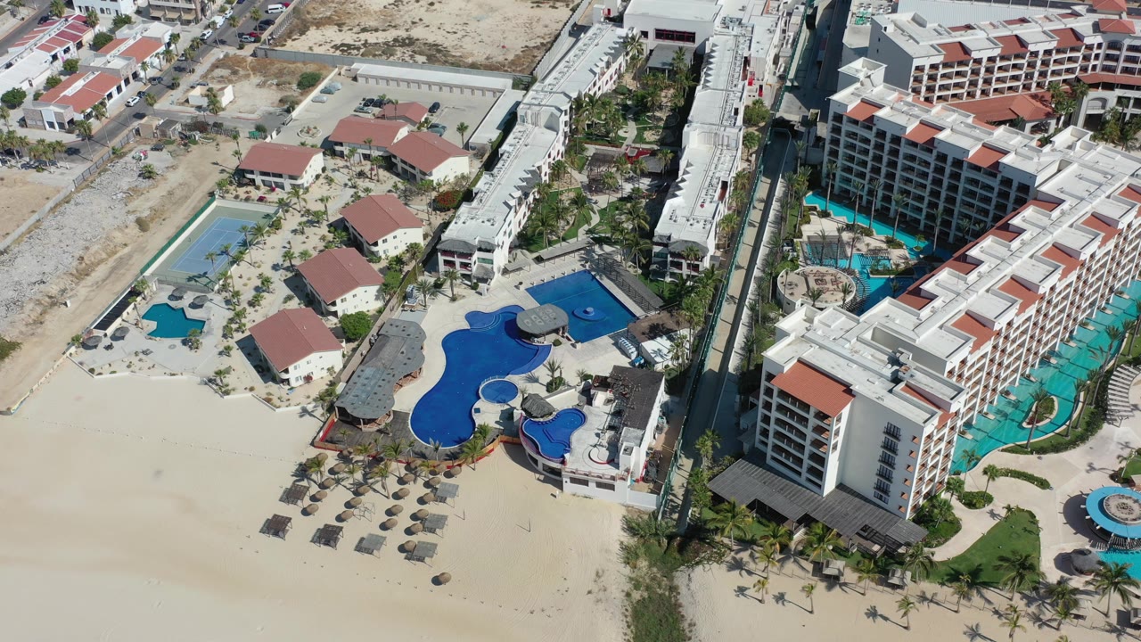 Aerial footage over Los Cabos, Mexico frames a large curved blue pool, smaller pools, white multilevel lodging, red roof sections, palm landscaping, and pale sand, with no visible people clearly identifiable.