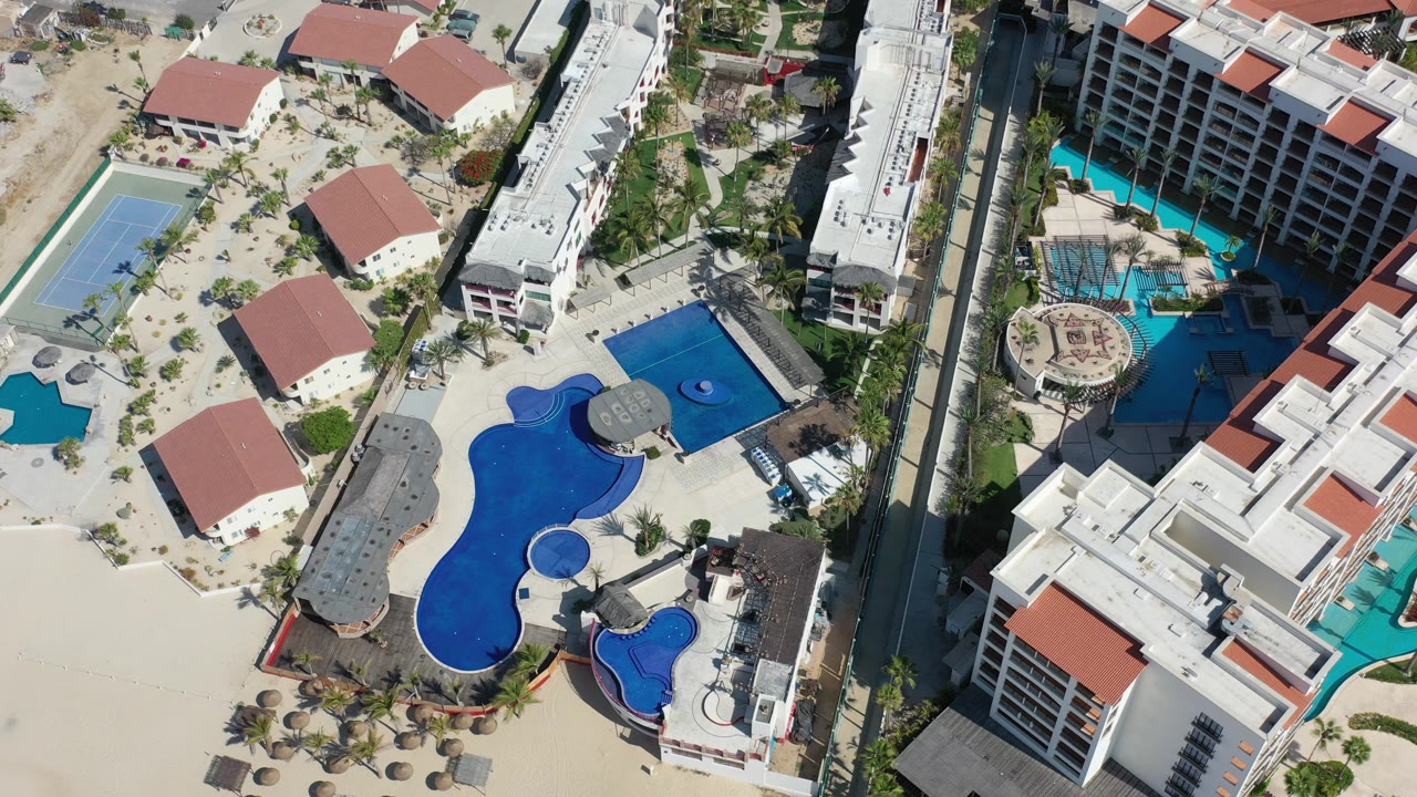 Aerial view over a Los Cabos resort area with blue pools, red-roofed buildings, pale sandy grounds, a road, and no visible people.