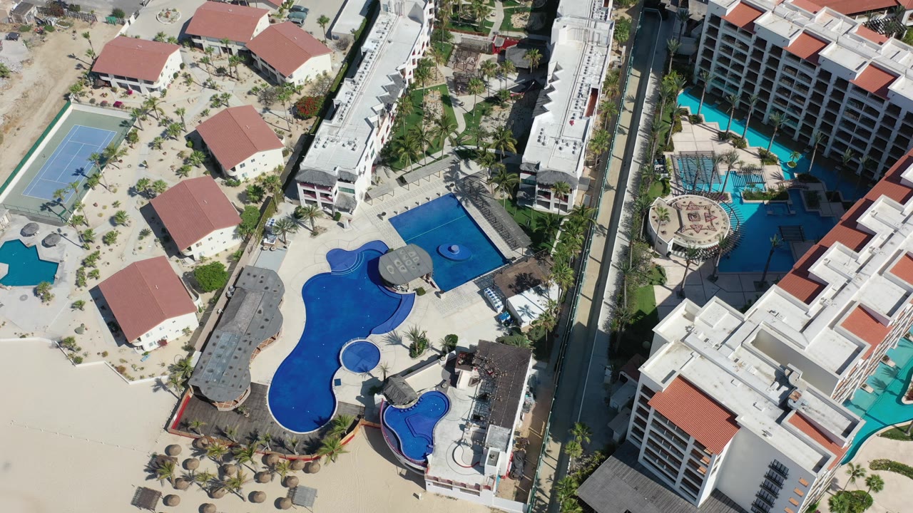 High overhead frame in Los Cabos showing curved blue pools, a circular pool feature, red roofs, hotel blocks, sandy walkways, and no visible people.