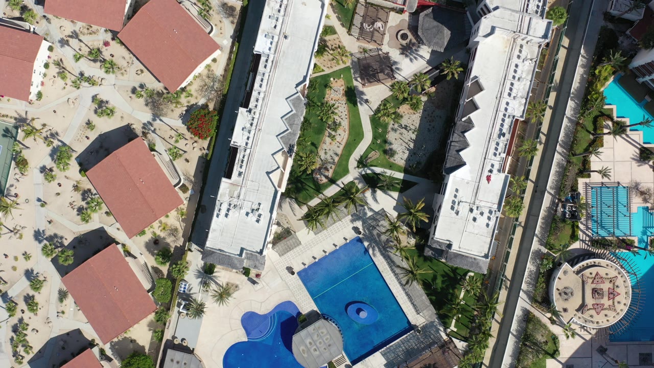 Top-down view of red-roof buildings, pools, palms, and a long white roof.