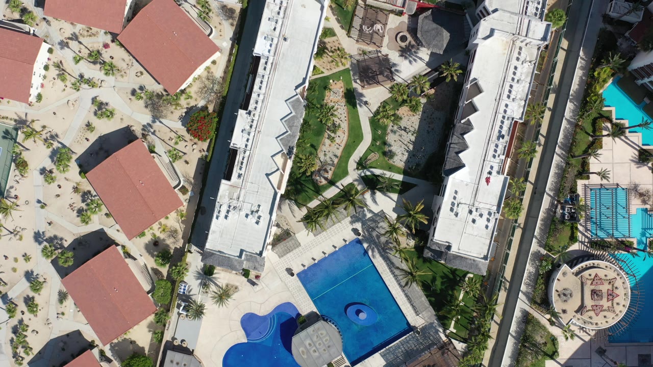 Los Cabos overhead view with red roofs at the left, two bright blue pools near the center, landscaped paths, and no visible people.