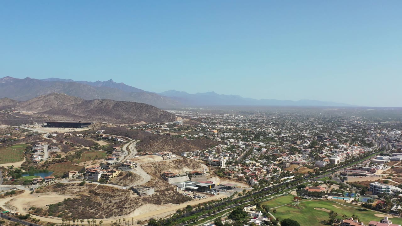 Aerial view of homes, roads, green lawns, dry hills, and distant mountains.