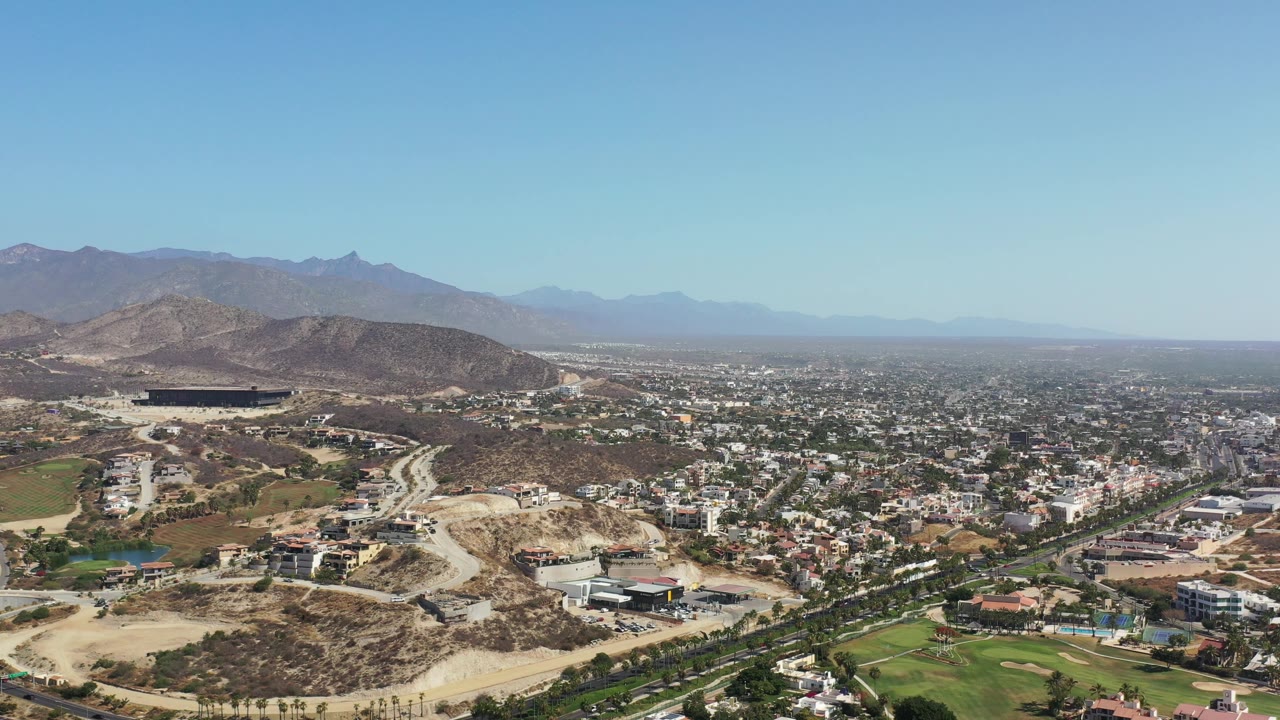 Second frame of the Los Cabos aerial with a hillside development area at left, dense housing to the right, green lawns below, distant mountains, clear sky, and no visible people.