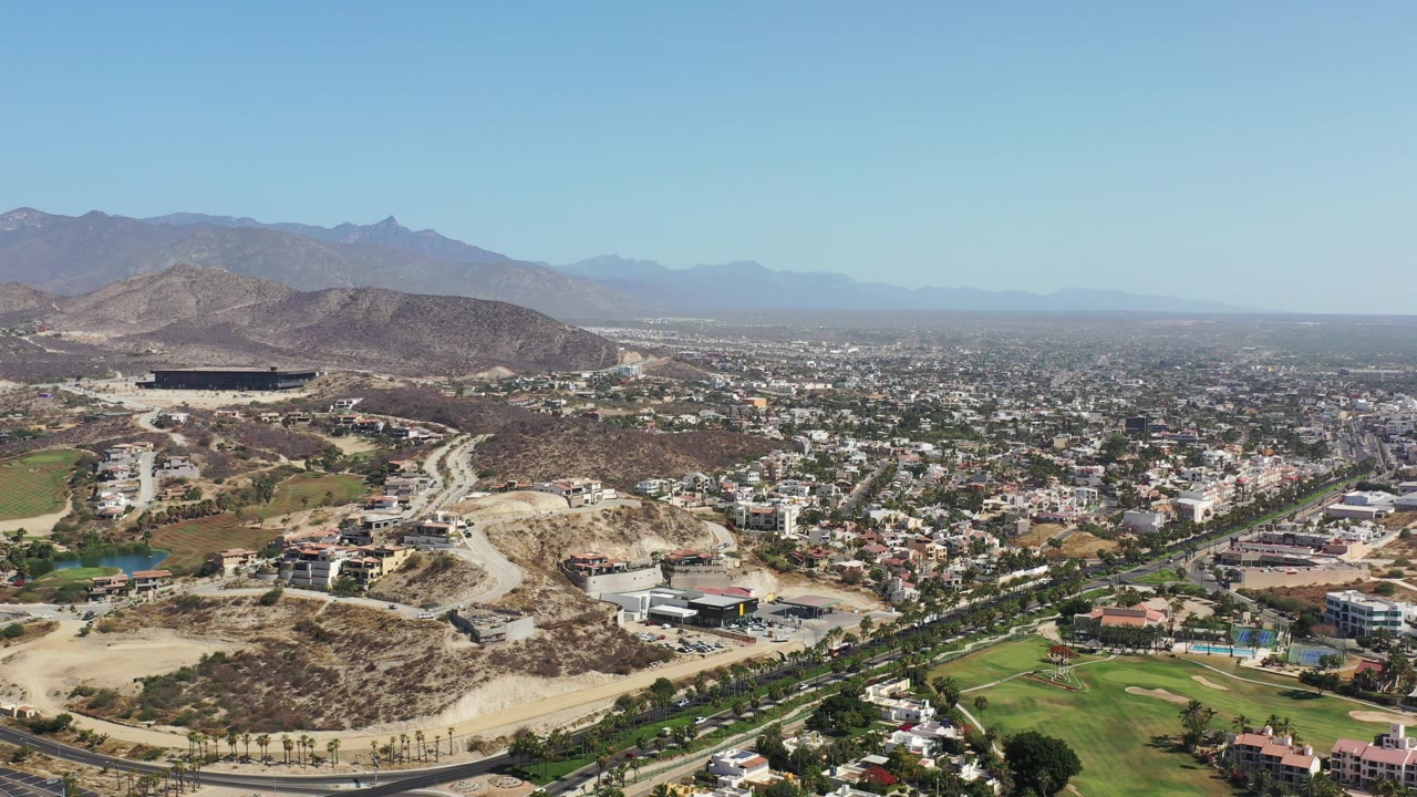 Wide aerial view in Los Cabos, Mexico showing a landscaped green area, curving roads, residential buildings, dry hills, and distant mountains under clear daylight, with no visible people.