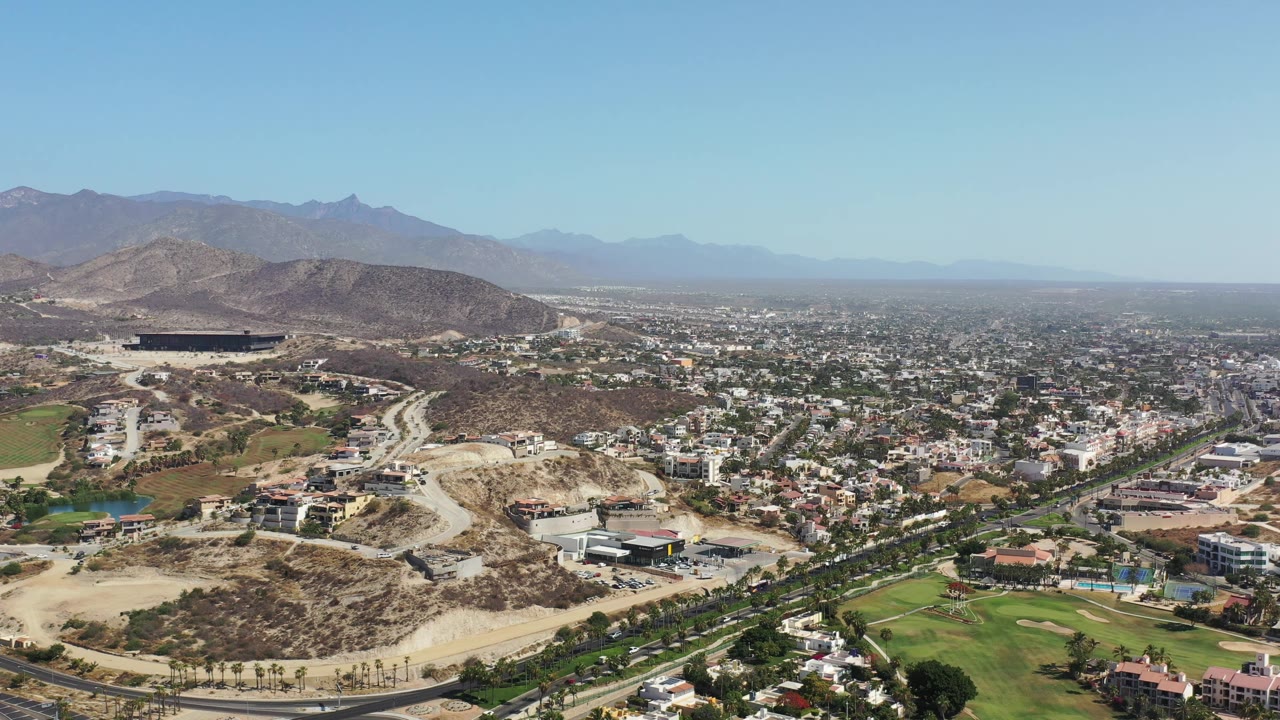High side-top view in Los Cabos, Mexico with winding roads crossing the foreground, clusters of light-colored buildings, green landscaping, dry hillside terrain, and no visible people.