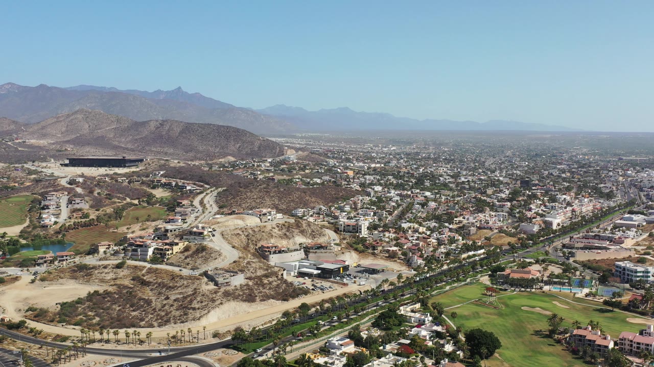 Drone-height town view with dry hills, roads, green lawns, and distant haze.