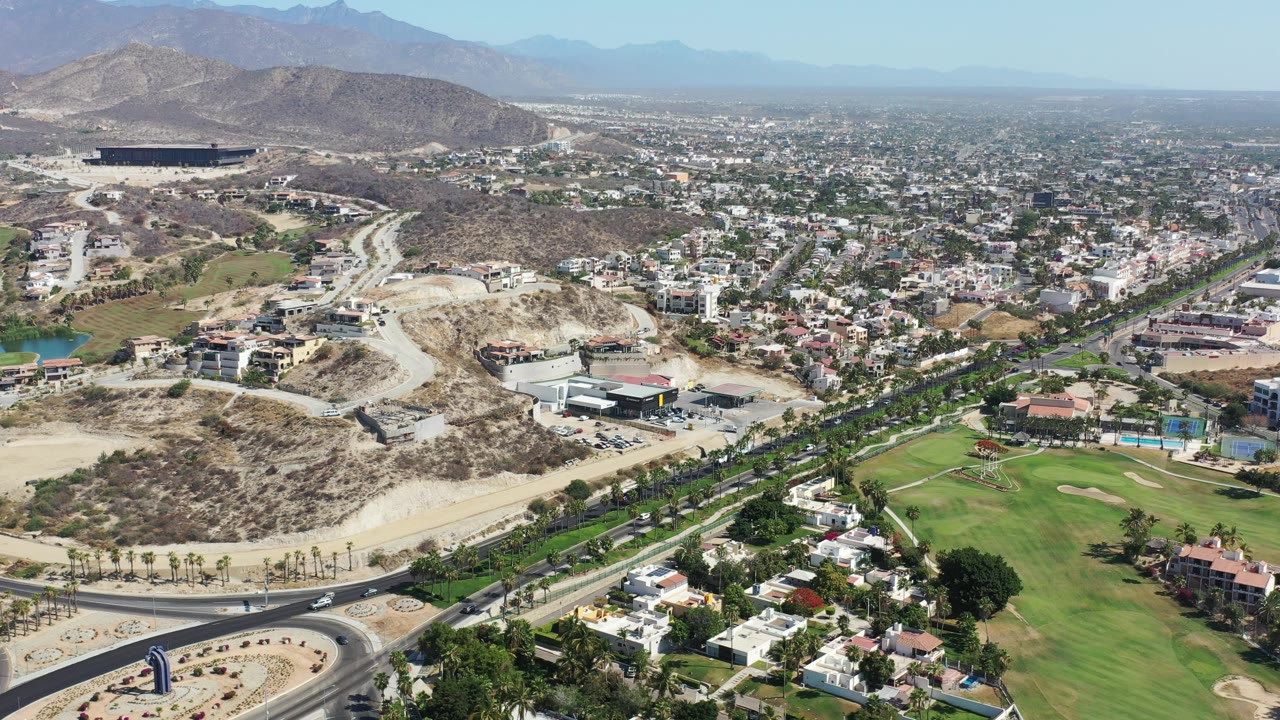 Aerial view over Los Cabos, Mexico, with a curved road in the foreground, green open space at right, hillside construction, dense housing, distant mountains, and no visible people.