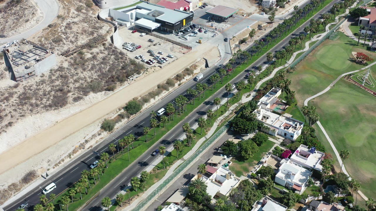 High aerial frame over Los Cabos, Mexico showing a highway with cars, white homes, palm-lined landscaping, green turf, and dry hillside, with no visible people.