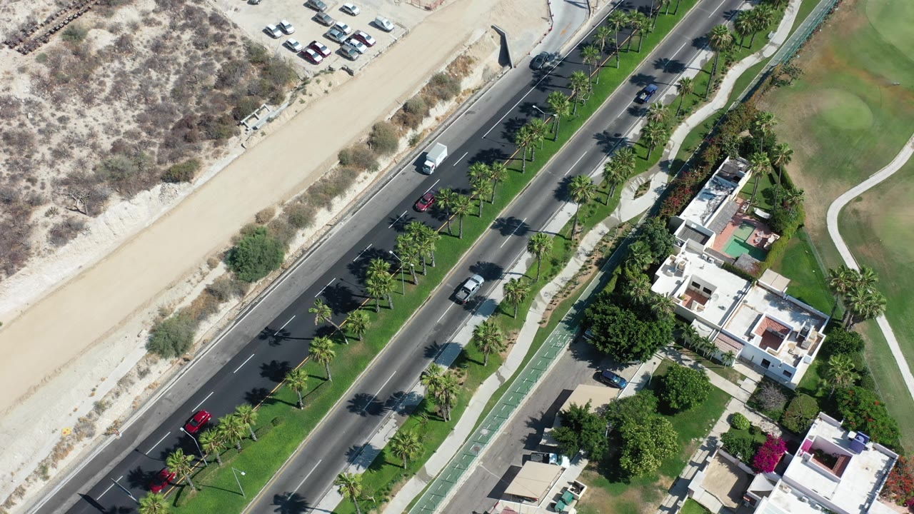 Overhead Los Cabos road view with vehicles moving past a sandy beach edge, palm-lined landscaping, and white residential buildings, with no visible people.