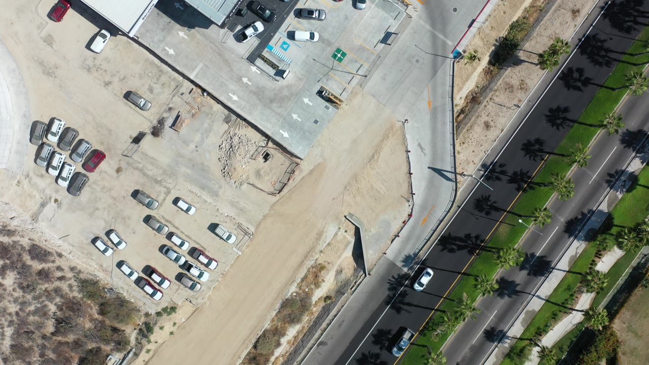 Top-down Los Cabos frame with a large pale roof, nearby parked cars, a curving road edge, sandy construction-like lots, and no visible people.