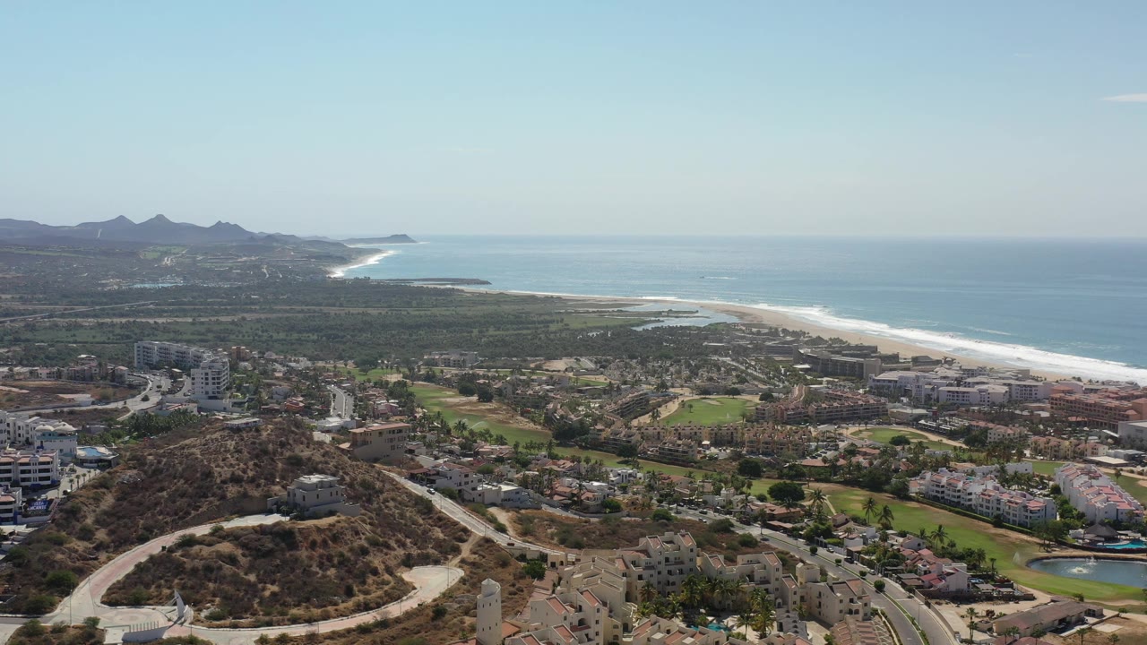 High aerial view over Los Cabos, Mexico, with dry hillside foreground, low buildings, green course areas, a long surf line, blue ocean, distant mountains, and no visible people.