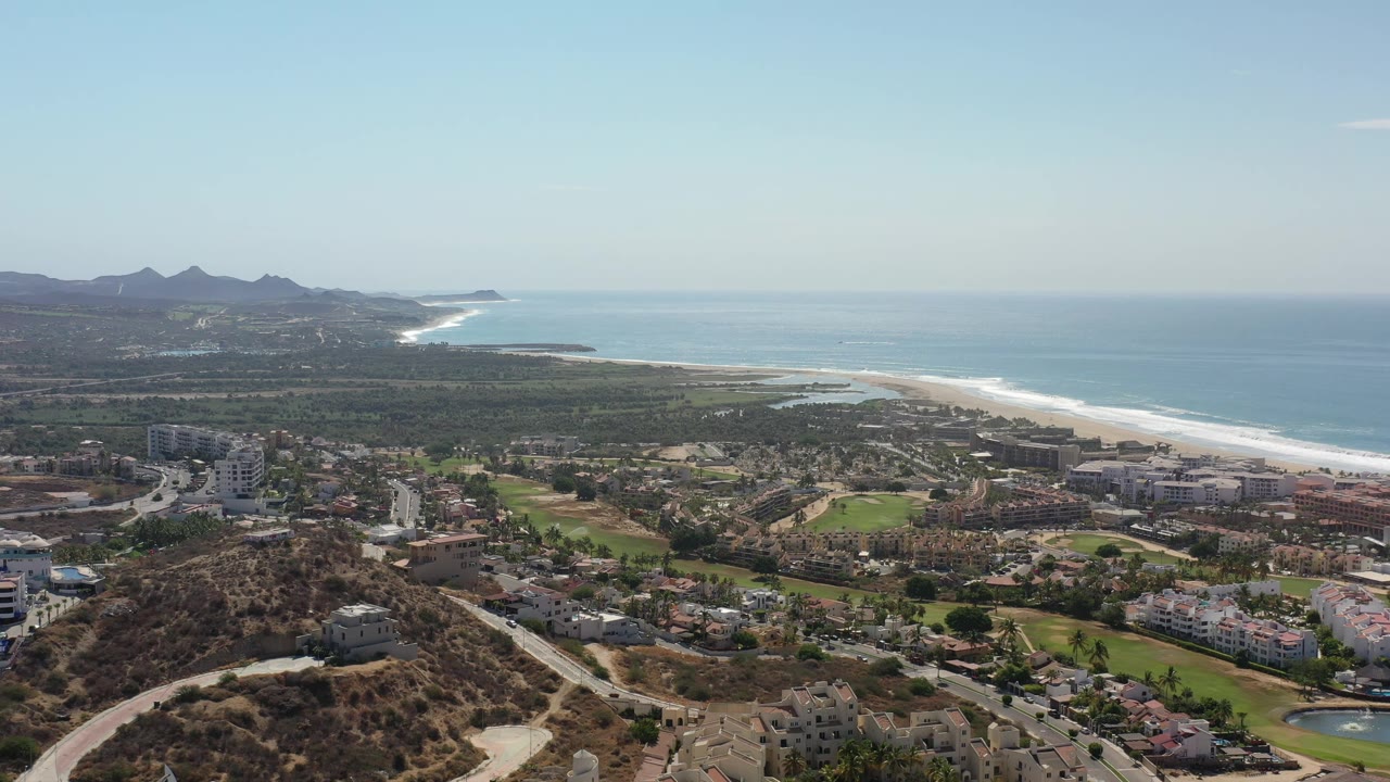 Los Cabos aerial view with residential blocks and green fairways below, white surf along the beach, blue water beyond, mountains on the horizon, and no visible people.