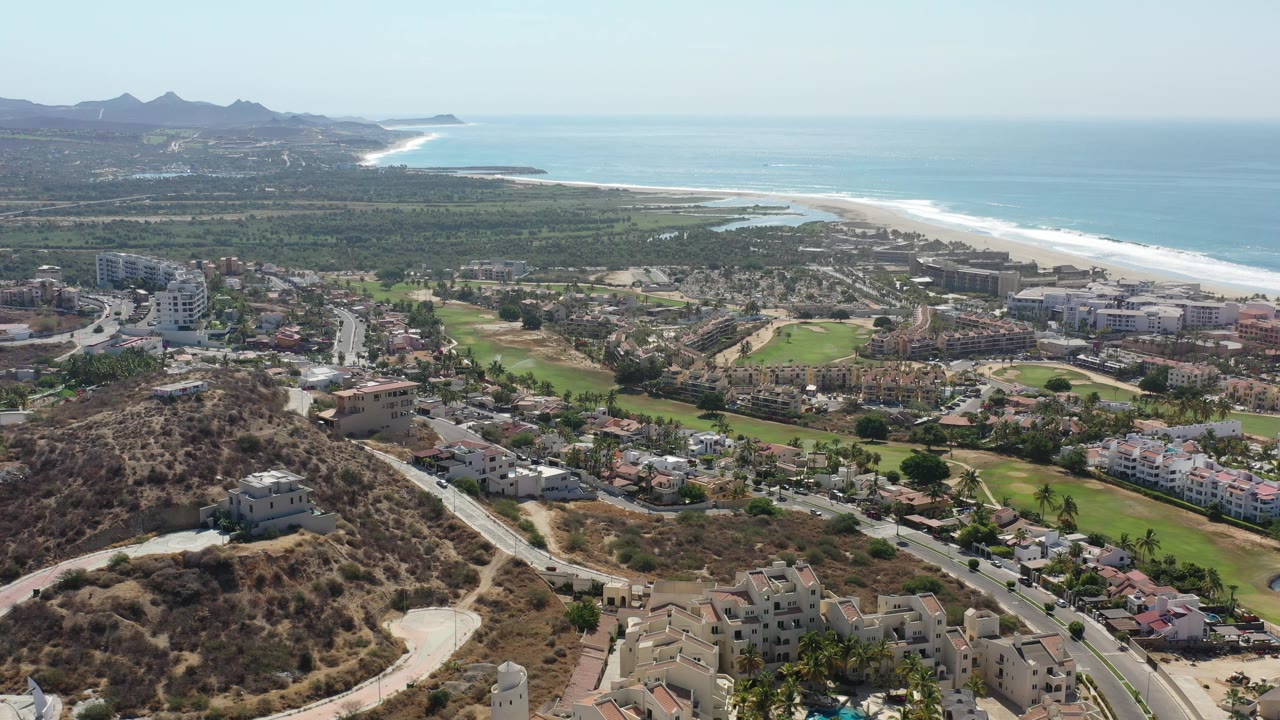 Aerial frame in Los Cabos, Mexico with a road network and hillside homes in front, green fairways and larger beachfront buildings beyond, ocean horizon, and no visible people.