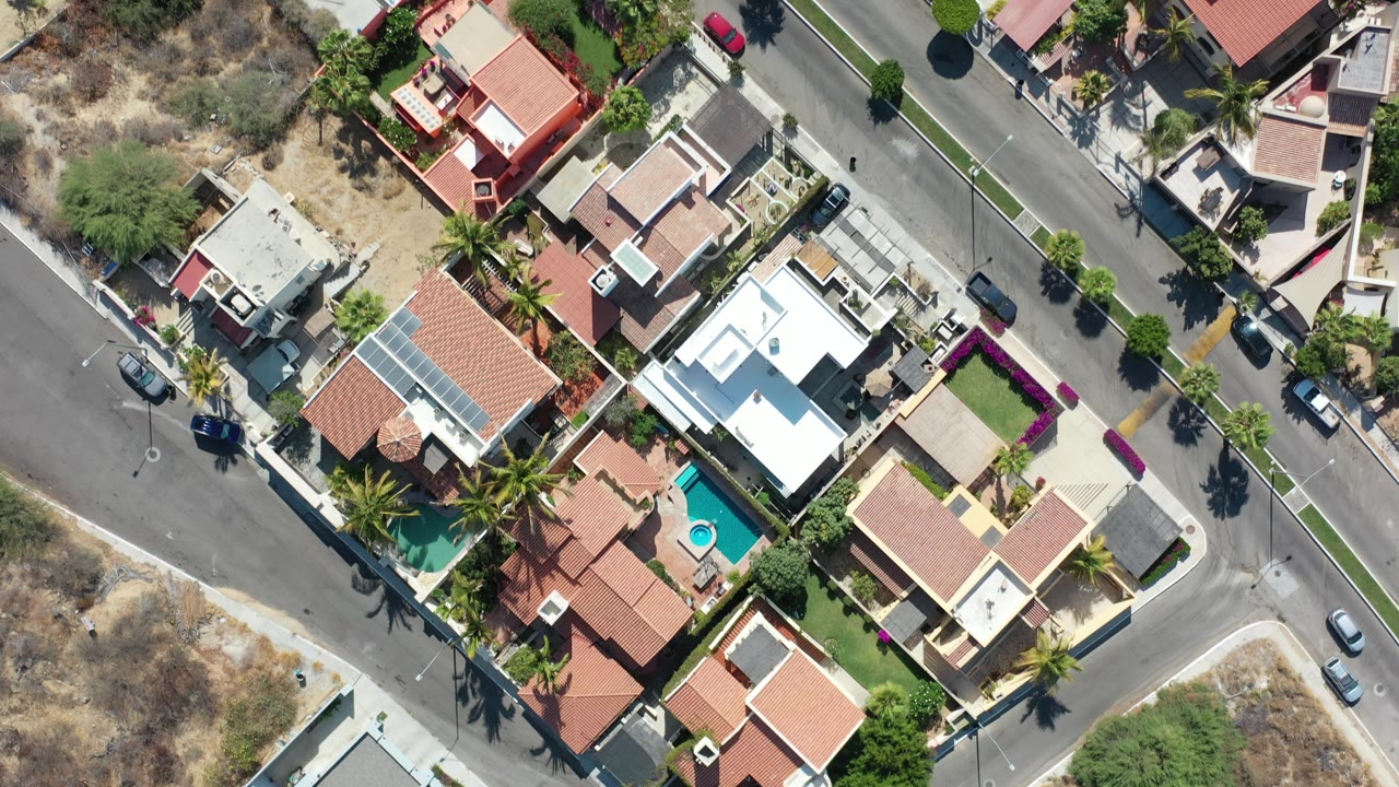 High overhead Los Cabos view showing a diagonal road between private homes, blue pools, palm-lined yards, parked cars, and no visible people.