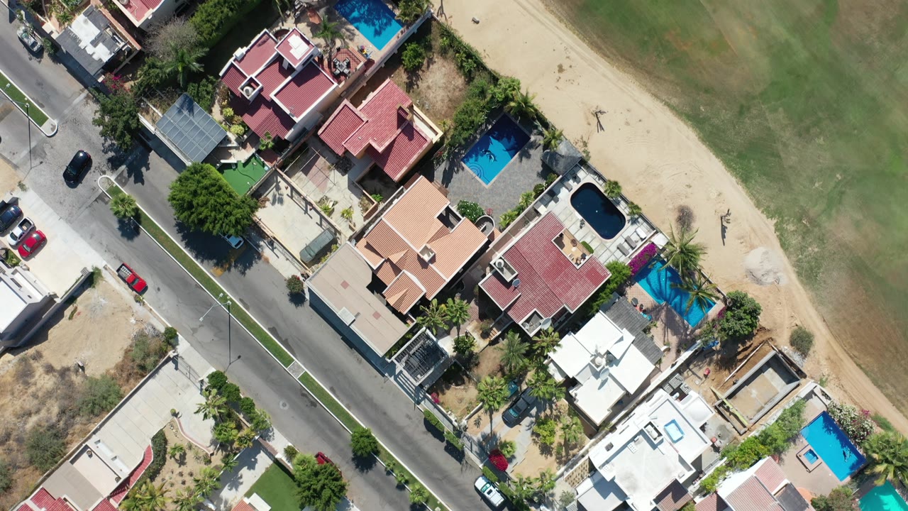 Aerial straight-down view in Los Cabos, Mexico with clustered rooftops and pools along a road, bordered by sandy ground and green field, with no visible people.