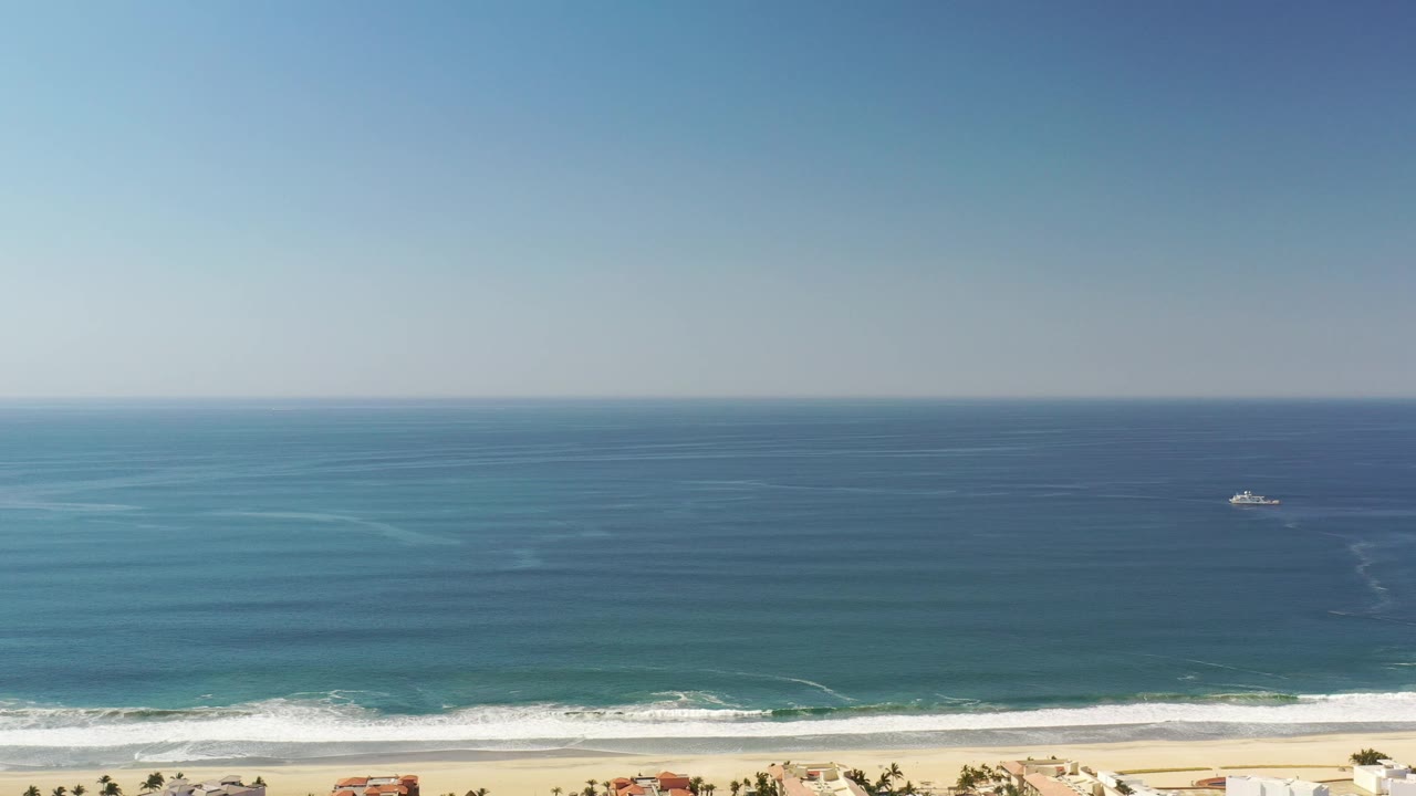 Opening frame of a Los Cabos, Mexico aerial view with blue ocean, a small offshore boat, white surf, sandy beach, coastal houses at the bottom edge, and no visible people.