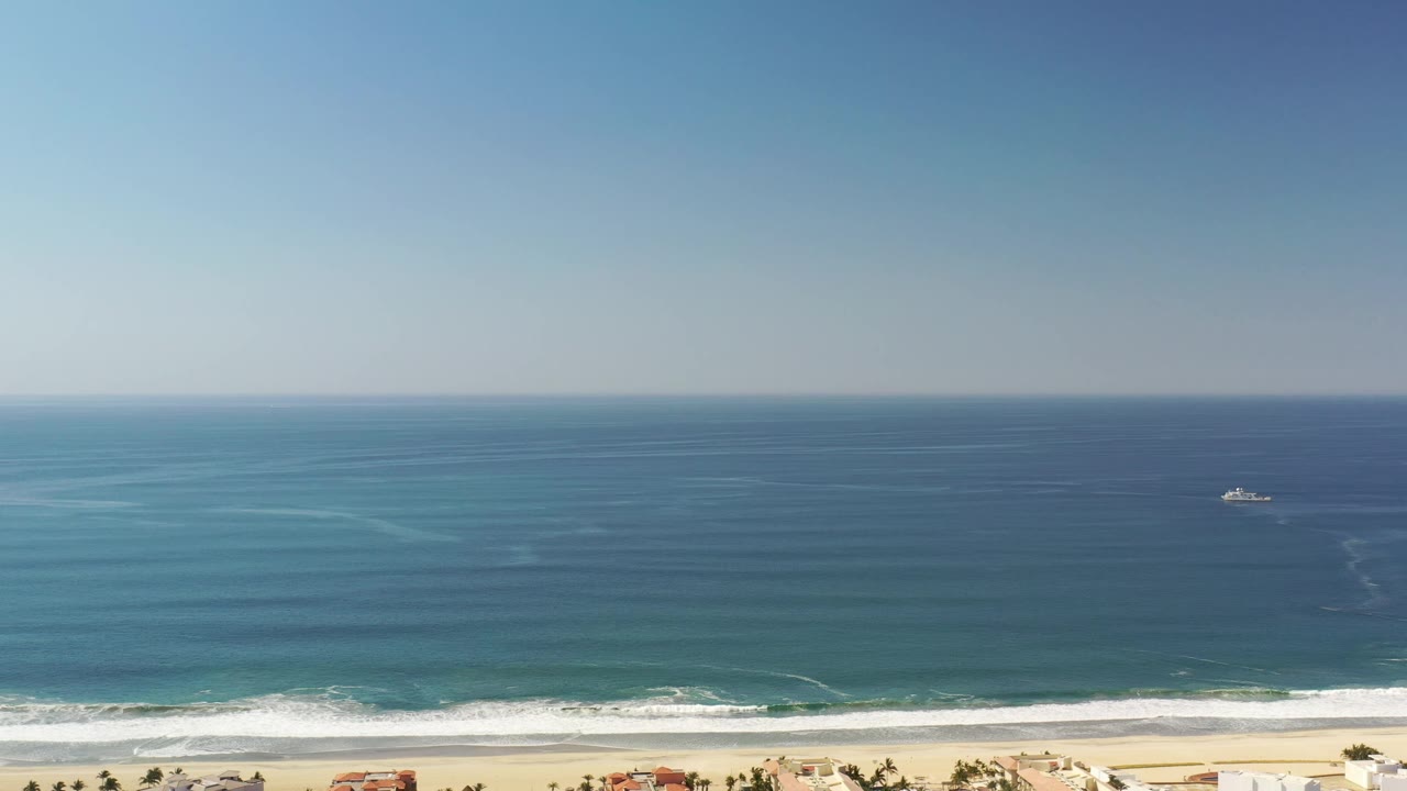 Los Cabos shoreline seen from above, with the ocean filling most of the frame, surf breaking near the sand, beach homes along the lower edge, and no visible people.