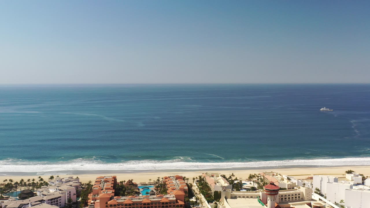 Opening aerial frame over the Los Cabos, Mexico coastline with deep blue ocean, a white surf line, pale sand, beachfront buildings, and no visible people.