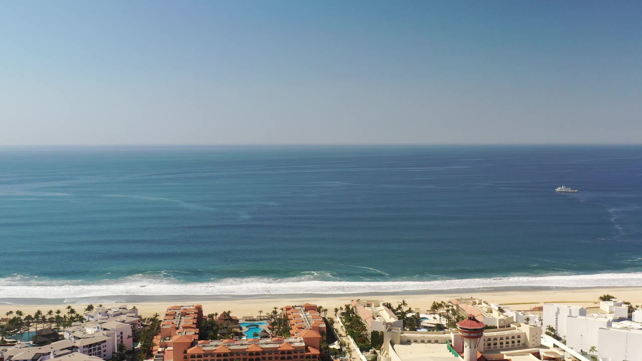 Aerial view in Los Cabos, Mexico showing turquoise water closer to shore, white waves, beachfront buildings, palms, and no visible people.