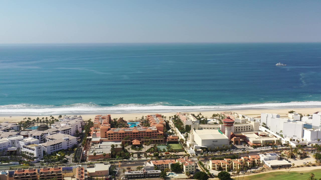 Aerial Los Cabos view with the ocean horizon high in frame, beach and surf through the center, white buildings and palms below, and no visible people.