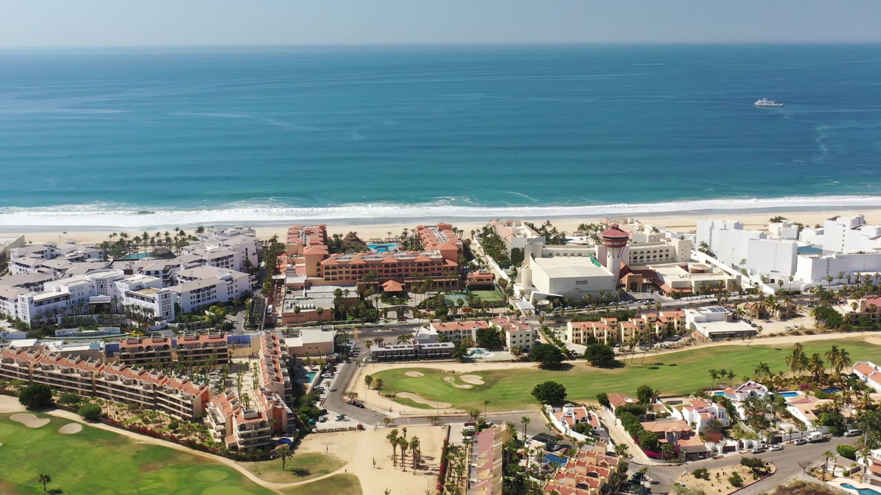 High aerial view over Los Cabos, Mexico, with turquoise ocean beyond a white surf line, beachfront buildings, golf-course greens, and no visible people.