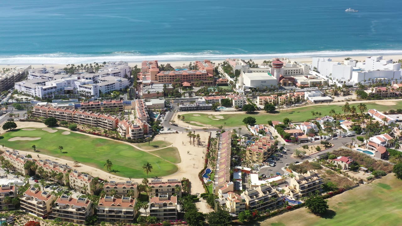 Los Cabos aerial view one second in, showing the central road and surrounding buildings more prominently, with golf greens below and beach surf stretching across the background.