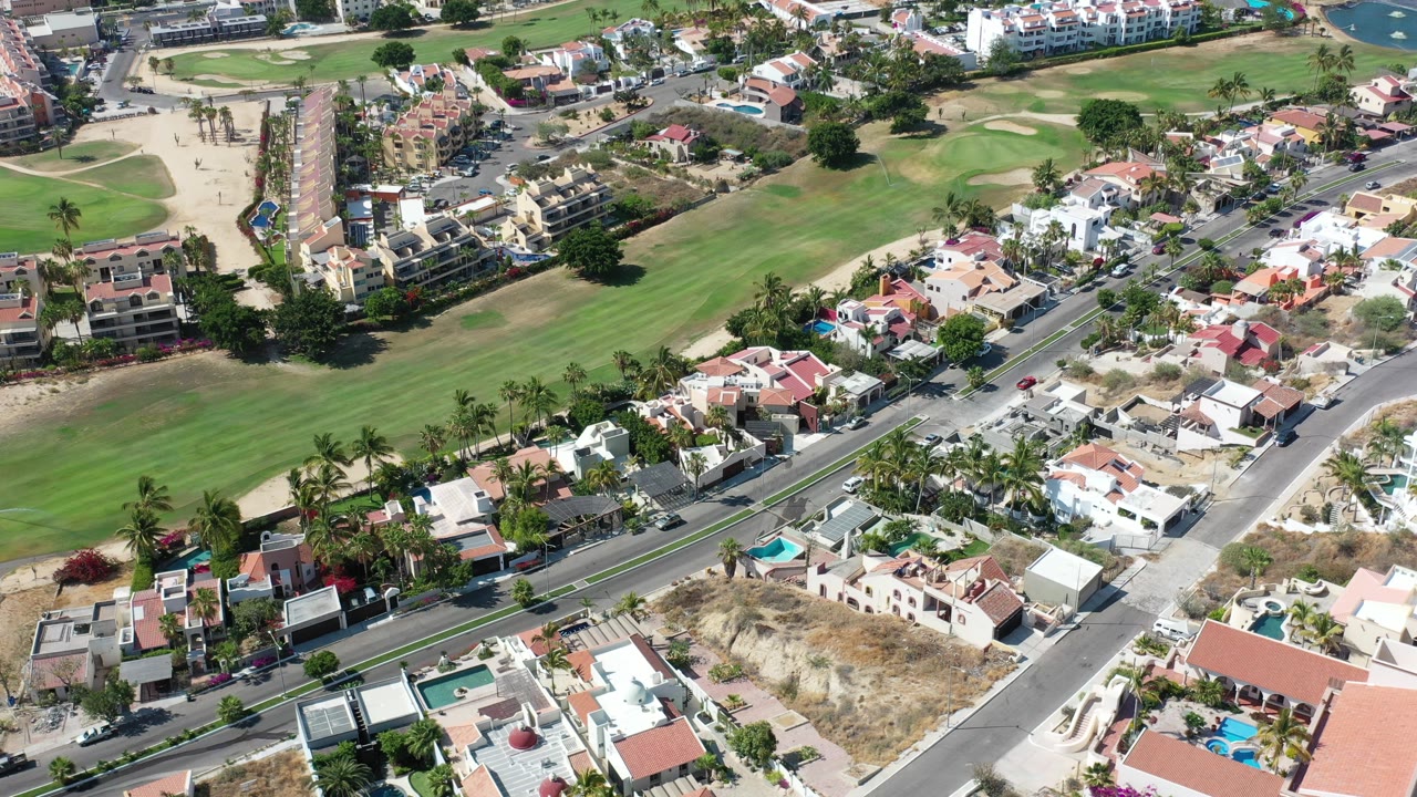 High aerial view over Los Cabos homes, a broad green open area, diagonal streets, palm trees, pools, and no visible people.