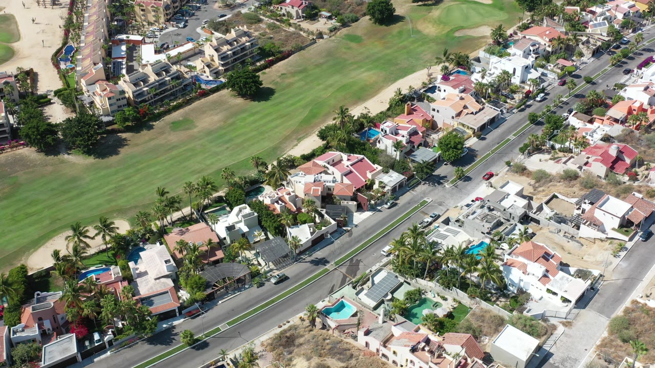 Opening aerial frame over Los Cabos, Mexico, showing a curving road beside homes, palm trees, blue pools, and a broad green fairway area with no visible people.