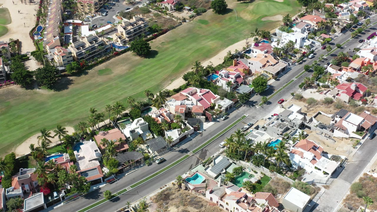 Aerial frame in Los Cabos, Mexico, with the road running diagonally between white and terracotta homes and golf-course grass, with no visible people.
