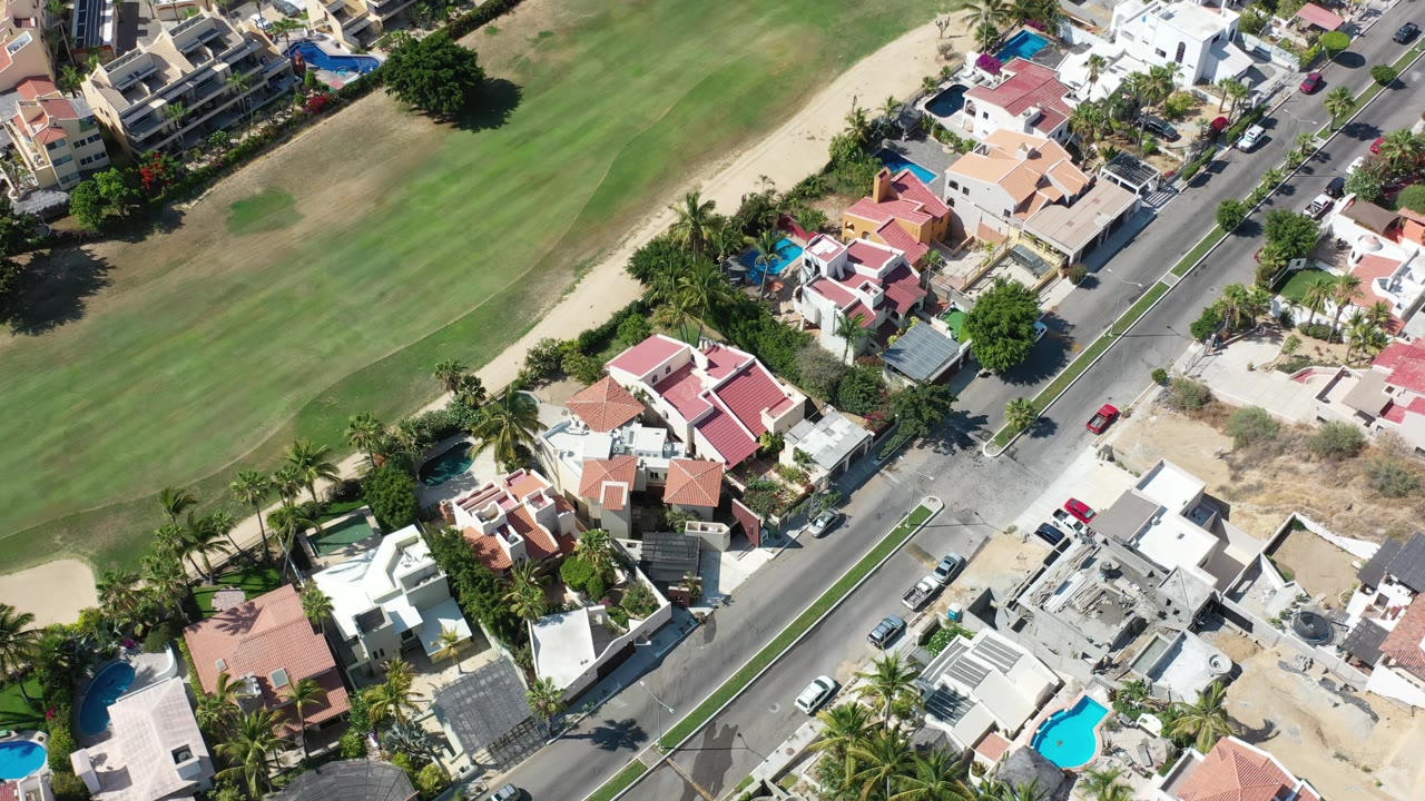 Aerial frame in Los Cabos, Mexico, with red-roofed and white homes beside a paved street, clipped edge of a green course, parked vehicles, palms, and no visible people.