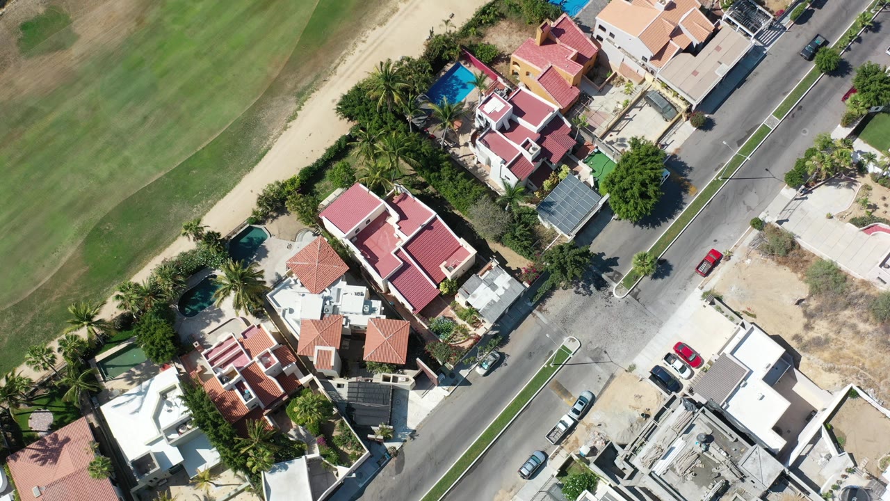 Overhead Los Cabos, Mexico frame showing red-roofed homes, palm trees, a road with cars, and a large green landscaped area, with no visible people.