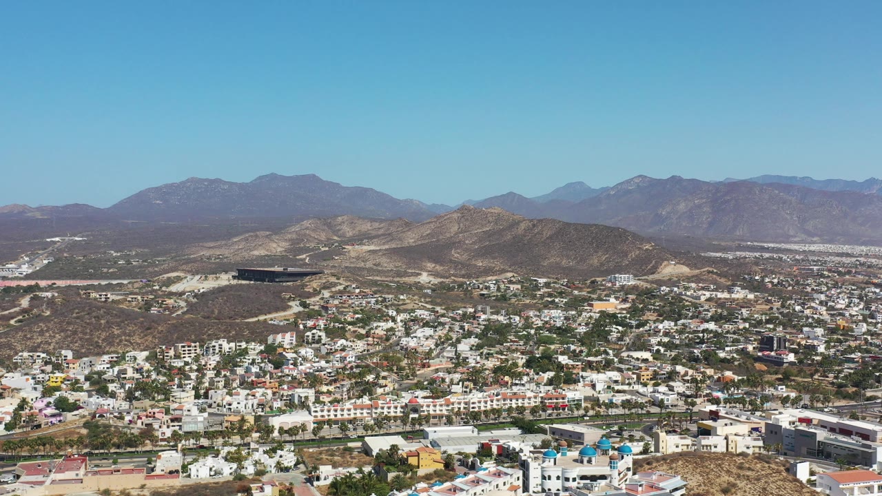 Opening aerial frame over Los Cabos, Mexico, showing white hillside buildings, arid ridges, a distant mountain range, blue sky, and no visible people.