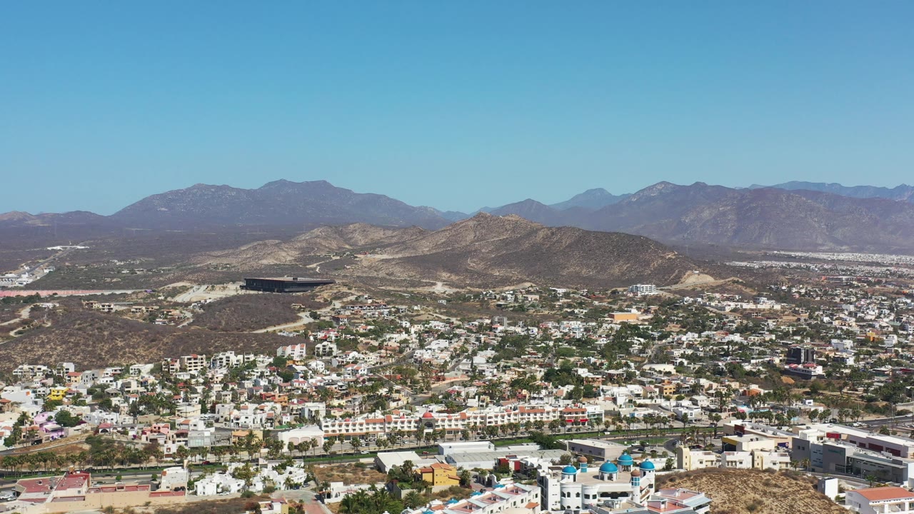 Los Cabos aerial view with dense white buildings in the lower frame, winding roads through dry hills, distant mountains, clear daylight, and no visible people.