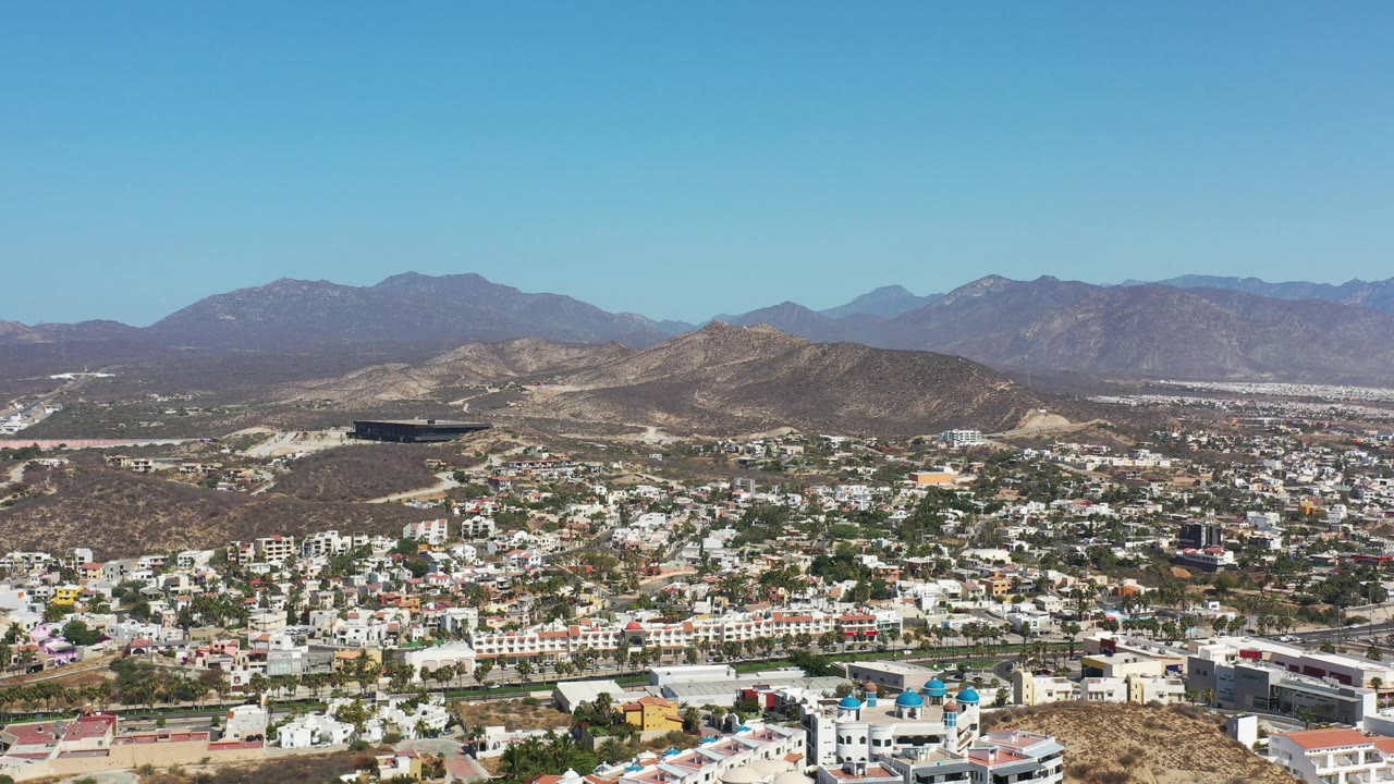 Aerial frame from Los Cabos, Mexico, with a hillside resort district, pale roads, scattered greenery, rugged brown hills, mountain horizon, and no visible people.