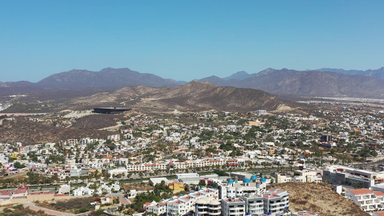 Wide aerial over dry hills, neighborhoods, resort buildings, and distant mountains.