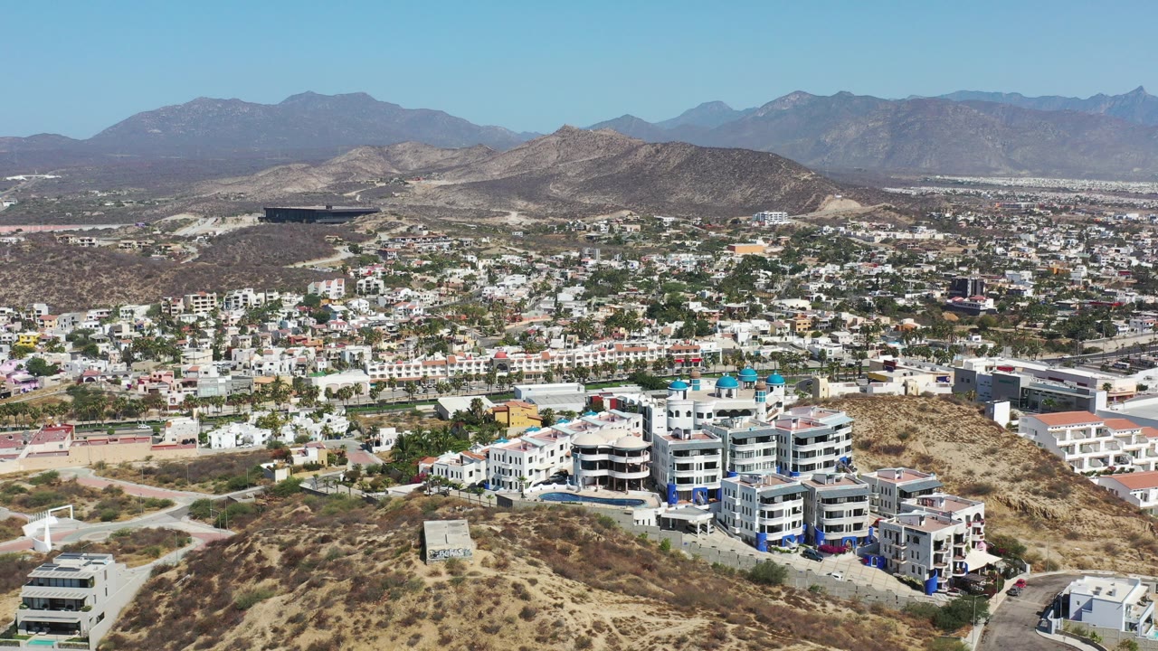 Wide aerial view of hillside buildings, city streets, dry hills, and distant mountains.