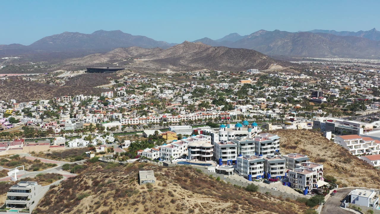 Second aerial frame over Los Cabos, Mexico, with a dry ridge in front, clustered white buildings below, a broad urban area in back, hazy mountains, and no visible people.