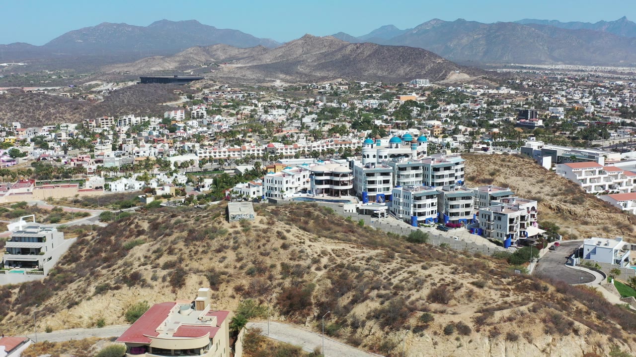 Opening aerial frame over Los Cabos, Mexico, with dry hillside foreground, white residential buildings in the midground, blue-gray mountains on the horizon, and no visible people.