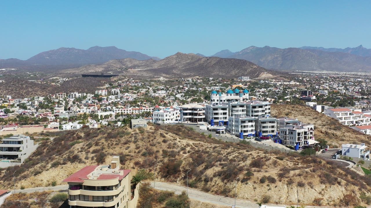 Aerial view over dry hills, residential buildings, city blocks, and distant mountains.