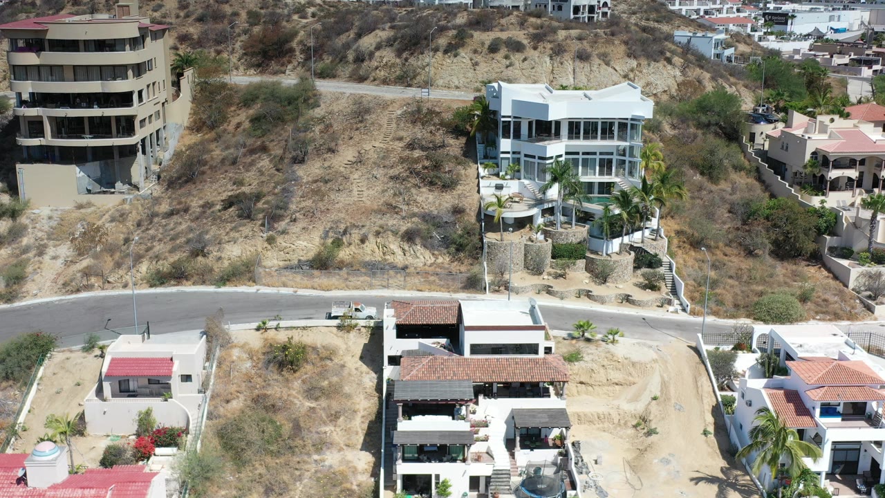 Los Cabos aerial frame showing modern hillside houses, tan scrub-covered slopes, a curving road, palm trees, and no visible people.