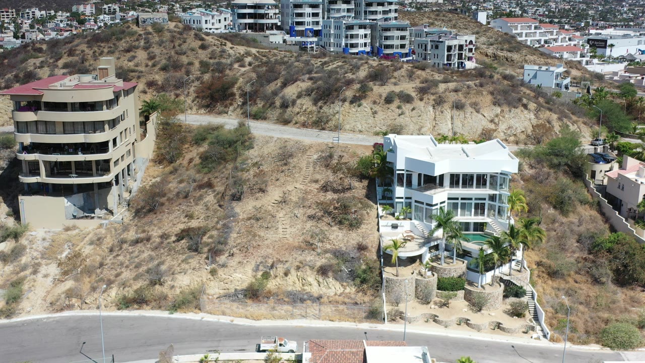 A Los Cabos hillside view shows a rounded tan apartment building, a white modern home, dry scrub terrain, a road, distant residences, and no visible people.